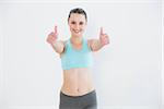 Portrait of a toned young woman gesturing thumbs up against wall in fitness studio