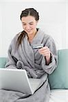 Casual young woman in bathrobe doing online shopping through laptop and credit card in bed