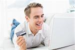 Cheerful casual young man doing online shopping through laptop and credit card on sofa