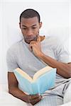 Relaxed young Afro man reading book in bed at house