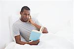 Relaxed young Afro man reading book in bed at house