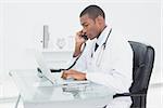 Side view of a concentrated male doctor using phone and laptop at medical office