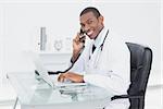 Side view portrait of a smiling male doctor using phone and laptop at medical office