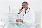 Portrait of a smiling male doctor using cellphone and laptop at medical office