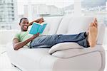 Full length of a smiling young Afro man reading a book on sofa at a bright house