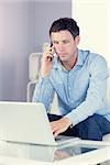 Attractive casual man using laptop and phoning in bright living room