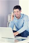 Smiling casual man using laptop and phoning in bright living room