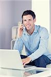 Cheerful casual man using laptop and phoning in bright living room