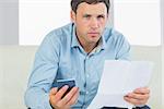 Serious casual man holding calculator and document in bright living room