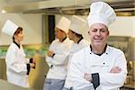 Mature male chef posing proudly in a kitchen with crossed arms and cooks in the background