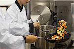 Two chefs working in a busy kitchen at the stove one tossing vegetables