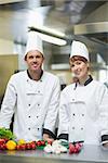 Two young chefs posing in a kitchen behind counter of vegetables