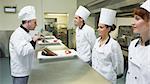 Three chefs presenting their plates to the head chef in a kitchen