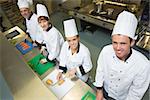 Four chefs preparing food at counter smiling at camea standing in a row in a kitchen