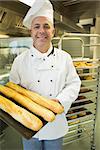 Mature baker presenting proudly some baguettes on a baking tray smiling at camera