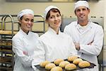 Three young bakers posing in a kitchen holding a baking tray with rolls on it