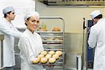 Cute young female baker holding a baking tray with rolls on it smiling at the camera