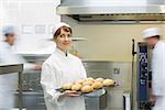 Cute female baker holding a baking tray with rolls on it smiling at the camera