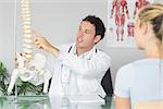 Handsome doctor showing a patient something on skeleton model in bright office