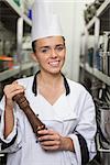 Young cheerful chef holding pepper mill between shelves in professional kitchen