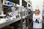 Young frowning chef standing arms crossed between shelves in professional kitchen