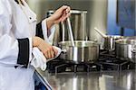 Young chef stirring with ladle holding pot in professional kitchen