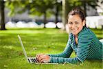 Attractive casual student lying on grass using laptop looking at camera on campus at college