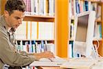 Handsome serious student using computer reading notes in library