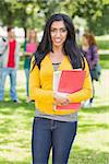 Portrait of college girl holding books with blurred students standing in the park