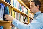 Side view of a smiling mature student selecting book from shelf in the library