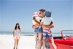 Father holding children on beach near convertible