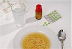 Medications, bowl of chicken soup and glass of water on table, studio shot