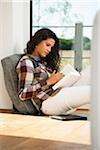 Teenage girl sitting on floor next to window and reading book, Germany