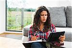 Teenage girl sitting on floor next to sofa, writing in binder and using tablet computer, Germany