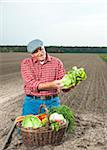 Farmer kneeling in field with basket of fresh vegetables, smiling and looking at lettuce, Hesse, Germany