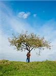 Farmer standing on hill next to apple tree, eating apple, Germany
