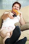 Unemployed man sitting on the couch in his underwear, watching TV and eating a sandwich.