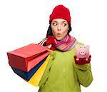 Concerned Mixed Race Woman Wearing Winter Clothes Holding Shopping Bags and Piggybank Isolated on White Background.