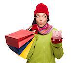 Concerned Expressive Mixed Race Woman Wearing Winter Clothes Holding Shopping Bags and Piggybank Isolated on White Background.