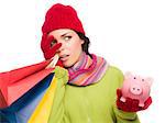 Concerned Expressive Mixed Race Woman Wearing Winter Clothing Holding Shopping Bags and Piggybank Isolated on White Background.