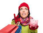 Stressed Mixed Race Woman Wearing Winter Clothing Looking Up Holding Shopping Bags and Piggybank Isolated on White Background.