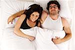 Beautiful couple laid smiling enjoying a lie-in in the morning in a white bed