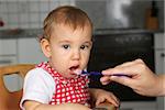 Baby opens mouth wide, mother feeding it with porridge in the kitchen