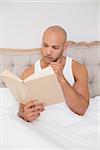 Relaxed young bald man reading book in bed at house