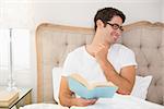 Smiling relaxed young man reading book in bed at house