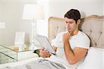 Casual young man reading newspaper in bed at home