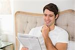 Casual smiling young man reading newspaper in bed at home