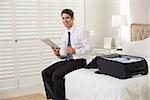 Smiling young businessman with coffee cup reading newspaper by luggage at a hotel room