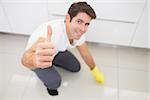 Portrait of a smiling young man cleaning the kitchen floor while gesturing thumbs up at house