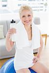 Smiling young woman with towel around neck and water bottle sitting on exercise ball